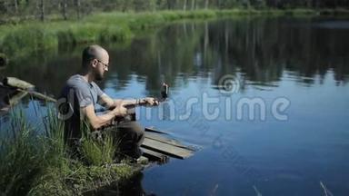 一个男人坐在湖边的码头上，用<strong>自拍</strong>棒在手机上做<strong>自拍</strong>。 美丽的蓝色湖泊和