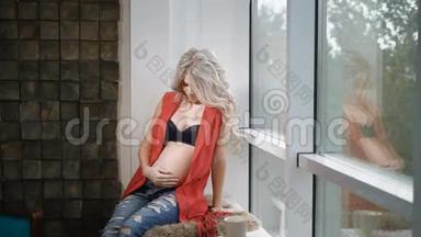 年轻的<strong>孕妇</strong>穿着红色衬衫，蓝色牛仔<strong>裤</strong>和黑色胸罩坐在靠近墙的窗台上。 未来母亲