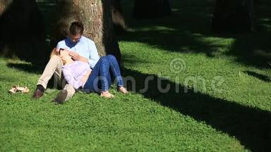 一对浪漫的情侣<strong>坐在</strong>棕榈<strong>树下</strong>。 一个女孩在一个男人的大腿上。 一对相爱的夫妇在草地上的公园里休息
