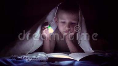 一个孩子晚上用手电筒在毯子下面读一本书。 有着淡发和蓝眼睛的男孩