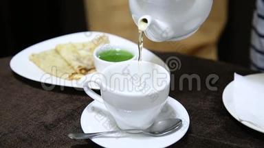 用瓷壶把茶倒入桌上的白杯中。 背景上的果冻和煎饼