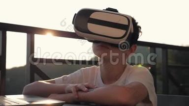 这个男孩正在电脑上玩虚拟现实眼镜在日落时。 现代概念