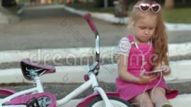 一个穿着粉红色裙子的小女孩坐在台阶上的公园里，戴着耳机享受着智能手机。
