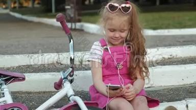 一个穿着粉<strong>红色</strong>裙子的小女孩坐在<strong>台阶</strong>上的公园里，戴着耳机享受着智能手机。