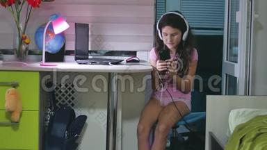 带着耳机坐着玩智能手机的少女。 女生在室内智能手机上玩网络游戏