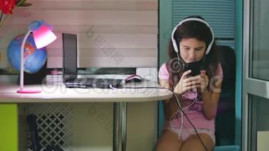 带着耳机坐着玩智能手机的少女。 在室内玩智能手机的女生