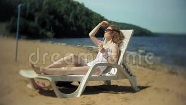 一个穿着白色比基尼的年轻女孩躺在海边沙滩上的休息室里晒太阳，喝着鸡尾酒