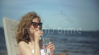 一个穿着白色比基尼的年轻女孩躺在<strong>海边</strong>沙滩上的休息室里<strong>晒太阳</strong>，喝着鸡尾酒