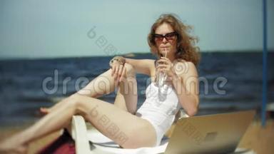 一个穿着白色比基尼的年轻女孩躺在<strong>海边</strong>沙滩上的休息室里<strong>晒太阳</strong>，喝着鸡尾酒