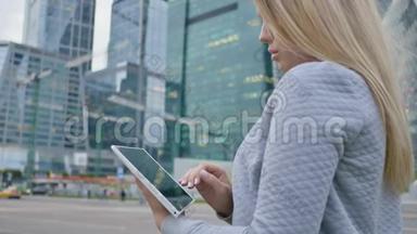 一个年轻的金发女孩在市中心摩天大楼的背景上使用平板电脑。