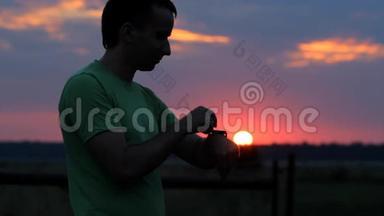 一个人在日落时看并触摸智能手表。 美丽的红天和树林后面的太阳
