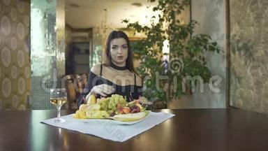 一个年轻的年轻的黑发女人，穿着黑色的裙子，乳沟，独自吃水果，葡萄，在高档餐厅的餐桌上。