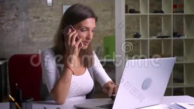 有魅力的女办公室工作人员在室内工作场所用电话和笔记本<strong>电脑打字</strong>