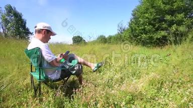 一个男人坐在折叠的野<strong>餐椅</strong>上，用智能手机工作。 周围是挥动的草，被风倾斜。