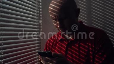 一个带智能手机的人站在窗户边带百叶窗