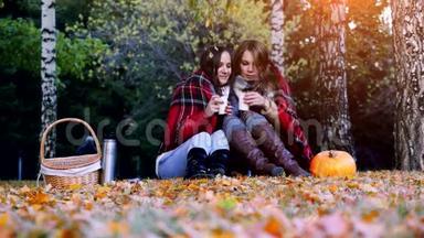 秋天公园里，年轻漂亮的女人坐在野餐的毯子里，喝着热水瓶里的热茶。 女孩地毯