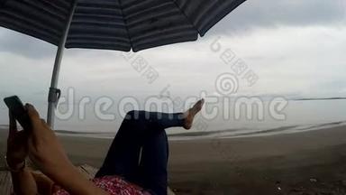 成熟的女人在热带海滩上躺在遮阳伞下，用移动智能手机浏览互联网