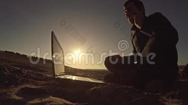 一个商人，自由职业者，打着电话，坐在沙滩上后面，坐在沙滩上，在阳光下自由职业者的剪影里