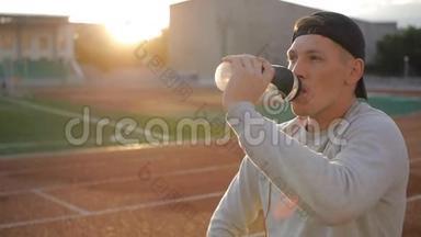 戴着naseball帽子的疲惫的运动员在<strong>体育场跑道</strong>上喝着瓶子里的水