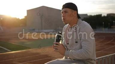 戴着naseball帽子的疲惫的运动员在体育场跑道上喝着瓶子里的水
