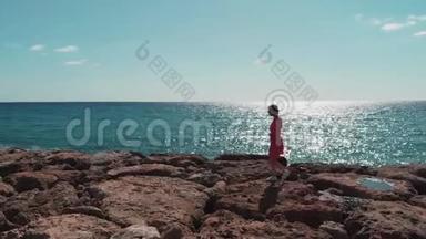 穿着红色衣服的女人走在岩石码头海滩上，海浪拍打着悬崖，阳光明媚的日子里阳光明媚。 太阳之路蓝色的倒影