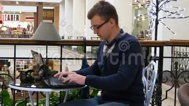 戴眼镜的年轻人坐在<strong>商场</strong>的桌子旁，用笔记本电脑工作