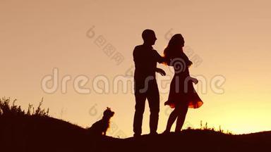 一对相爱的情侣在日落时跳舞剪影和接吻。 爱的男人和女人与狗跳舞剪影自然慢