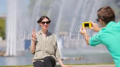 男朋友坐在喷泉的背景下给他的朋友拍照。 年轻人在女人的照片上