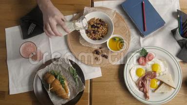 咖啡厅健康早餐的顶级景观。 人在格兰诺拉浇牛奶，英式早餐：煎鸡蛋，培根吐司在盘子里