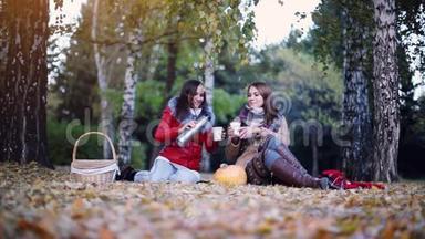 年轻的女人坐在野餐上，在秋天公园的<strong>热水</strong>瓶里<strong>喝</strong>热茶。 女孩们围在南瓜旁边