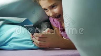 十几岁的女孩和夜狗在毯子下玩智能手机。 睡前的小女孩躺在床上冲浪