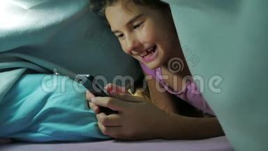 少女之夜和狗在毯子下玩智能手机。 <strong>睡前</strong>的小女孩躺在床上冲浪