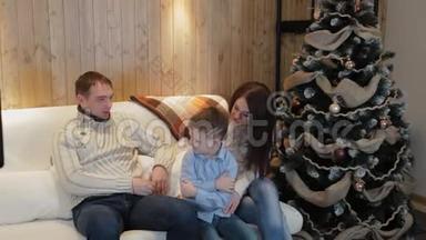 美丽幸福的三口之家在沙发上微笑着，在<strong>圣诞夜</strong>的圣诞树旁交谈。