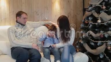 美丽幸福的三口之家在沙发上微笑着，在圣诞夜的圣诞树旁交谈。