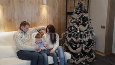美丽<strong>幸福</strong>的三口<strong>之家</strong>在沙发上微笑着，在圣诞夜的圣诞树旁交谈。