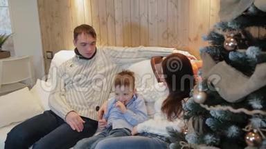 美丽幸福的三口之家在沙发上微笑着，在圣诞夜的圣诞树旁交谈。