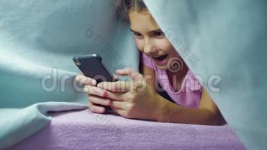 女孩少年和狗在一个夜毯下<strong>玩</strong>智能<strong>手机</strong>。 睡前的小女孩躺在<strong>床上</strong>冲浪