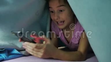 少女和狗在毯子下玩智能手机的夜晚。睡前的小女孩躺在床上冲浪