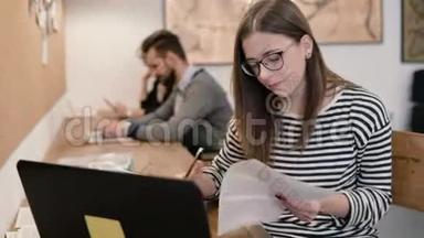 年轻漂亮的女孩正在使用笔记本电脑，处理文件，在现代的创业办公室做笔记