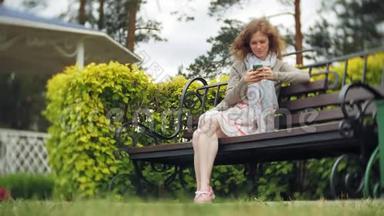 在<strong>美丽</strong>的绿色公园的长凳上使用智能手机放松的女人。 <strong>校园</strong>中的年轻千禧妇女做手势