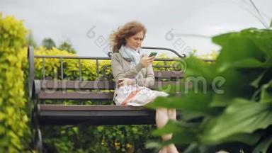 在美丽的绿色公园的长凳上使用智能手机放松的女人。 校园中的年轻千禧妇女做手势