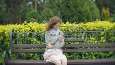 在美丽的绿色公园的长凳上使用智能手机放松的女人。 校园中的年轻千禧妇女做手势
