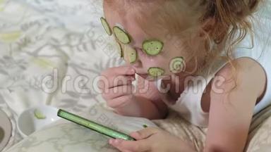 一个漂亮的小女孩兴高采烈地在家里<strong>制作</strong>黄瓜面具，并使用智能<strong>手机</strong>