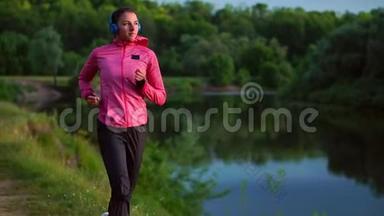 戴着长头发耳机的布鲁内特早上在公园里沿着河边跑步，夏天的日出时穿着粉红色的衣服