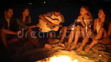 日落海滩派对，篝火晚会。 朋友们围坐在篝火旁，喝着啤酒，对着吉他唱歌。 青年