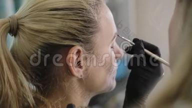 化妆师用专业的画笔在美容院的镜子前给一个漂亮的女孩画眉毛。