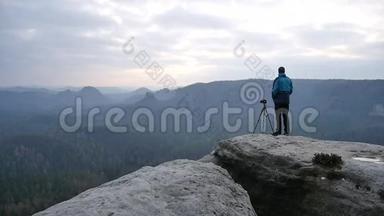 穿着蓝色夹克衫的摄影师站在岩石的视点上，拍摄朦胧的早晨景观。 在岩石里远足。