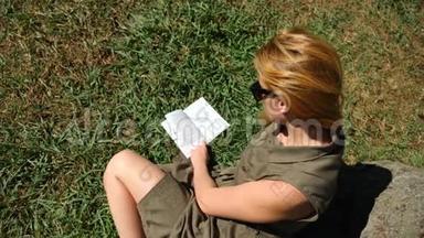 金发女人在公园里看书，<strong>坐在</strong>草地上，<strong>树下</strong>。 4k，慢镜头