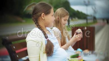 两个小女孩，苗条又胖，吃健康有害的食物，蔬菜和蛋糕坐在咖啡馆的长凳上，