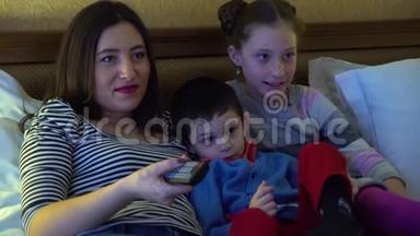 一位年轻的母亲带着两个孩子，躺在旅馆的床上看电视。 使用电视遥控器。 他们很开心。